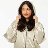 Campera cortavientos de acabado metalizado color dorado, con capucha ajustable mediante cordones, cierre frontal de cremallera negra y puños elásticos.