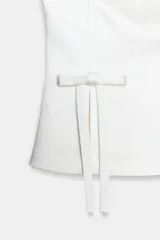 Top strapless de satén color blanco, con diseño de escote palabra de honor y hombros descubiertos. Presenta detalle de dos lazos decorativos en el frente y aberturas en el bajo. Cierre posterior con cremallera metálica.