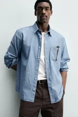 Camisa de corte regular fit confeccionada en tejido chambray de algodón con efecto lavado. Presenta cuello con botones, manga larga con puños abotonados, bolsillo en el pecho y cierre frontal de botones.