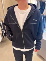 Campera con capucha negra de corte boxy, con cierre frontal completo, bolsillos tipo canguro y detalles de líneas blancas horizontales y en las mangas. Cuenta con un pequeño logo bordado en el pecho.