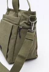 Cartera tipo tote bag de nylon color verde militar, con diseño amplio y liviano. Cuenta con asas superiores reforzadas, correa larga regulable y desmontable, bolsillos frontales y laterales con cierre metálico, y un pequeño aplique con logo en la parte inferior.