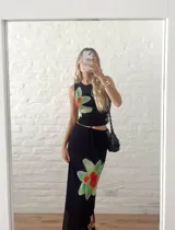 Conjunto de dos piezas compuesto por un top sin mangas y una pollera larga, confeccionados en microtul negro con estampado floral en tonos verdes y rojos. El top es ajustado y la pollera presenta un diseño de doble capa con cintura elástica.