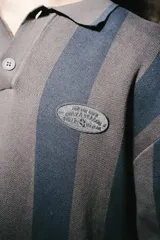 Chomba de manga larga con diseño de rayas verticales en tonos marrón y azul. Presenta cuello polo con cierre de botones y parches decorativos en el pecho.