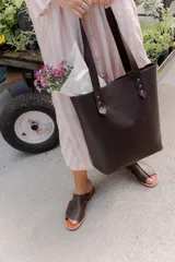 Cartera tipo tote bag de cuero marrón, con diseño minimalista, asas largas y detalles de remaches metálicos en la unión de las asas.