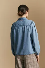 Camisa de denim azul con cuello camisero, cierre frontal de botones y mangas largas abullonadas con hombros fruncidos.