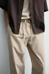 Pantalón relaxed fit color beige con rayas verticales finas, confeccionado en tejido de algodón. Cuenta con cintura elástica ajustable con cordón, bolsillos frontales y bolsillos traseros de plastrón.