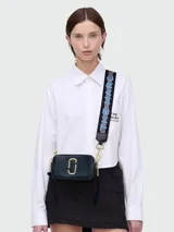 Cartera bandolera Marc Jacobs modelo The Snapshot, color azul con detalles dorados. Presenta doble compartimento con cierre, logo de la marca en el frente y correa de tela con el nombre de la marca.
