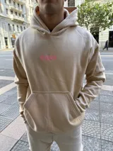 Canguro beige con capucha, de corte oversize. Presenta un diseño estampado en la espalda con la palabra 'Amore' en letras blancas grandes sobre un fondo de texto repetitivo en color rosado.