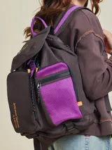 Mochila marrón con detalles en violeta. Tiene un bolsillo frontal superior con cierre, un bolsillo lateral de red y el logo de Rusty Worldwide estampado en naranja.