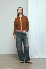 Cardigan de punto color marrón, con cuello vuelto, cierre frontal de botones y detalles de ribetes a rayas en los puños, la cintura y la botonera. Las mangas presentan un diseño tejido en relieve con textura de ochos.