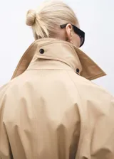 Abrigo tipo trench color beige, de corte oversize y largo hasta la rodilla. Presenta cuello con solapa, cierre frontal cruzado con doble botonadura, cinturón desmontable, dos bolsillos laterales y mangas largas con puños abotonados.