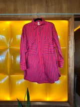 Camisa de corte oversize con diseño de rayas verticales en color rojo y azul. Presenta cuello clásico, cierre frontal con botones, mangas largas con puños abotonados y dos bolsillos frontales con solapa. El diseño tiene un largo asimétrico, siendo más corto en la parte delantera y más largo en la parte trasera.