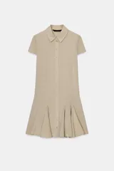 Vestido corto tipo camisero color beige, con cuello solapa, manga corta y cierre frontal con botones a presión. Presenta un diseño con pliegues en el bajo que le otorgan volumen.