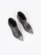 Bota de caña baja color negro con efecto gastado, confeccionada en cuero vacuno. Presenta punta fina, taco fino, y tira con hebilla y dije de corazón colgante alrededor del tobillo.