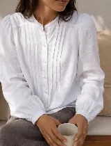 Camisa blanca de corte clásico con cuello mao y cierre frontal de botones. Presenta mangas largas con puños elásticos y detalles de tablas verticales en el frente.