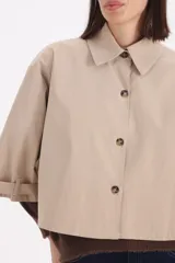 Piloto corto estilo camisa en color beige, con cuello clásico, cierre frontal de botones y mangas anchas con detalle de trabas en los puños.
