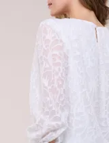 Blusa blanca de organza con textura devoré floral, silueta semiholgada y forro interno. Tiene escote bote, mangas largas abullonadas con transparencias, puños anchos con botones y ruedo elastizado.