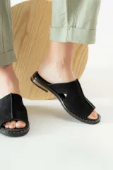 Sandalias tipo mule de gamuza negra con diseño de puntera abierta y costuras centrales visibles. Presentan una suela plana con detalle de tachas metálicas en el borde y una franja de madera en el talón.