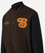 Campera bomber de estilo college con cuerpo a cuadros en tonos marrones y mangas largas de cuero sintético negro. Presenta cuello, puños y cintura acanalados a rayas, cierre abotonado, bolsillos laterales y aplique bordado de letra "B" en el pecho.