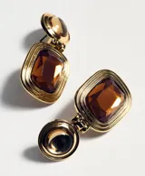 Pendientes colgantes dorados con diseño articulado, compuesto por una pieza superior circular abombada y un colgante rectangular inferior con una piedra de cristal marrón facetada y marco texturizado con líneas concéntricas.