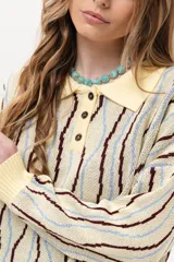 Buzo de punto tipo polo en tono amarillo claro con diseño de ondas a rayas en tonos marrón y celeste. Presenta cuello clásico, cierre frontal parcial con botones y silueta relajada con mangas largas.