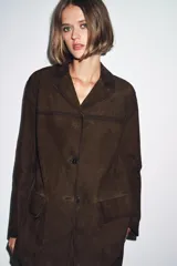 Blazer de mujer color marrón oscuro, confeccionado en piel de ante 100%. Presenta cuello solapa de muescas, manga larga, bolsillos de plastrón frontales y cierre frontal de botones.