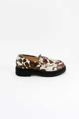 Mocasines estilo Mary Jane con diseño de estampado de vaca (cow print), confeccionados en cuero. Presentan una tira con hebilla ajustable sobre el empeine y una suela track negra de 4,5 cm de altura.