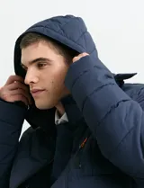 Campera acolchada de color gris, con capucha desmontable, cierre frontal y bolsillos laterales con cremallera. Presenta un diseño de costuras horizontales y detalles en color naranja en los tiradores de los cierres.