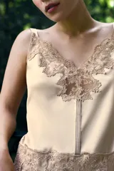 Vestido mini de tejido satinado color beige, con escote en pico adornado con encaje, tirantes anchos y detalle de encaje bordado en la cintura. El bajo presenta un acabado con volantes.