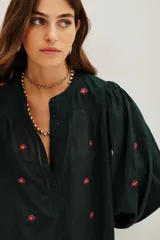 Camisa de algodón color crudo con bordados florales pequeños en azul marino. Presenta mangas largas abullonadas, cuello redondo y una abertura frontal con botones y detalles de encaje calado en la pechera y hombros.