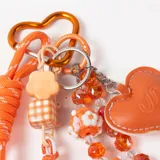 Llavero decorativo en tonos naranja, compuesto por una cuerda trenzada, un mosquetón en forma de corazón y múltiples dijes colgantes que incluyen cuentas de cristal, figuras de flores, un dije de corazón de cuero sintético y una esfera brillante.