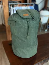 Mochila verde militar de lona con cierre superior con solapa y broche metálico. Tiene un bolsillo lateral con botón a presión y etiqueta de cuero con logo de la marca.
