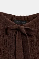 Jersey de punto calado color marrón, con cuello redondo ajustable mediante lazada y mangas largas con puños ajustables con lazos.