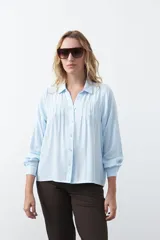 Camisa de manga larga en color celeste, con diseño de corte relajado, cuello clásico y detalle de tablas verticales en el frente.