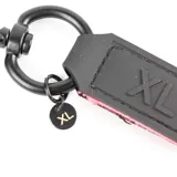 Llavero de cinta fucsia con herrajes metálicos negros, mosquetón superior y detalle de etiqueta con logo grabado.