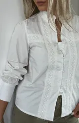 Camisa negra con cuello alto, mangas largas abullonadas y cierre frontal con botones. Presenta detalles de encaje y lentejuelas en el frente.