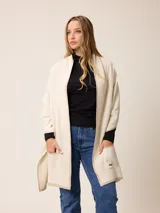 Poncho abierto de tejido de punto en color gris claro con bordes y detalles de bolsillos en gris oscuro. Presenta un diseño holgado y versátil con placa metálica decorativa en uno de los bolsillos frontales.