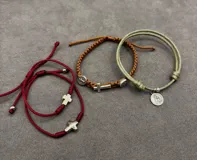 Set de tres pulseras ajustables de cordón con detalles religiosos. Incluye una pulsera roja con cruces, una pulsera marrón trenzada con cruz y medallas, y una pulsera verde con medalla colgante.