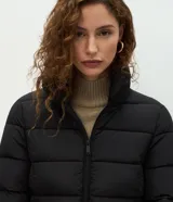 Campera puffer acolchada de color negro con pespuntes horizontales, cuello alto, cierre frontal y cinturón integrado ajustable para entallar la silueta.