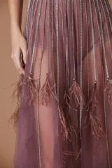 Vestido largo de tul color malva, compuesto por un body con escote en V y aplicaciones de tiras brillantes, cubierto por una capa transparente de tul con detalles de plumas en los puños y el ruedo.