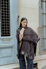 Ruana de tejido de punto grueso color negro, con diseño de pequeños cuadrados blancos y líneas rojas y azules. Tiene flecos largos en el borde inferior.