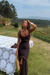 Vestido largo color chocolate, ajustado al cuerpo, con tirantes finos y escote en V. Presenta detalles de flecos en el escote y en la abertura lateral de la falda.