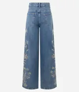 Pantalón wide leg de denim azul con bordados florales en los laterales. Presenta cintura baja, cierre con botón y cremallera, y bolsillos funcionales.
