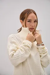 Sweater de punto color crudo, con cuello alto y cierre frontal parcial. Presenta un diseño de tejido con trenzas en las mangas y textura acanalada en el cuerpo.