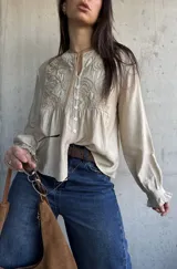 Camisa beige de manga larga con detalles bordados en el frente, cuello redondo y cierre frontal con botones. Las mangas presentan puños con volados.