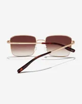 Gafas de sol Hawkers unisex modelo Iris, con montura cuadrada dorada de metal y lentes degradadas de marrón a transparente.