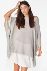 Blusa de punto estilo poncho en color beige, con diseño amplio, cuello redondo y aplique de etiqueta con el logo de la marca en el borde inferior.