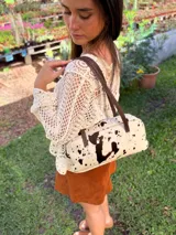 Cartera baguette de cuero color crudo con manchas negras, forro interno de tela y correa de cuero marrón.