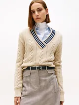 Sweater de punto color beige, con cuello en V ribeteado con franjas azul marino y blanco. Presenta tejido cableado (trenzas) en el frente y mangas largas.
