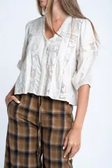 Blusa color crudo de manga corta abullonada, con escote en V y detalles de encaje y bordados verticales en el frente y mangas.